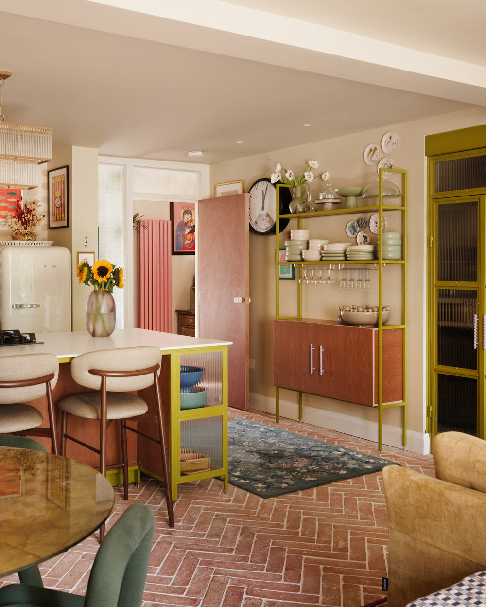 Olive green kitchen island with open shelving and reeded glass, alongside a bespoke mid-century style dresser displaying cocktail glasses and ornaments, finished with Sapele wood, a terracotta floor, and a Smeg fridge.