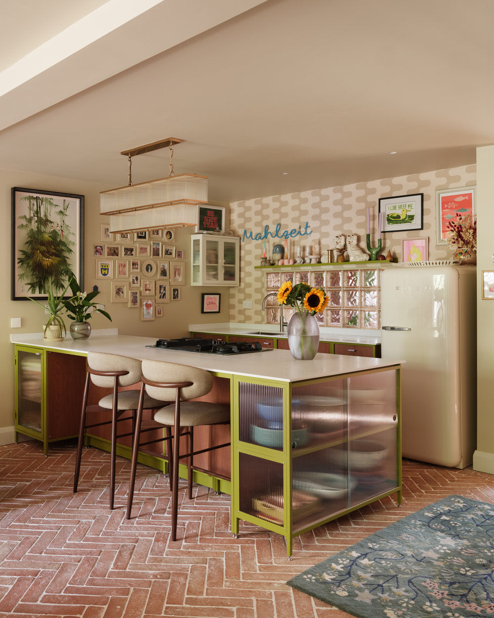Olive green steel frame kitchen with Sapele fronts and a terracotta floor, centred on a kitchen island with a hob and double-ended reeded glass cabinets for napkins, tablecloths, and plates. A chandelier hangs above, with glass bricks, a hatch, a Smeg fridge, and retro kitsch ornaments completing the space.