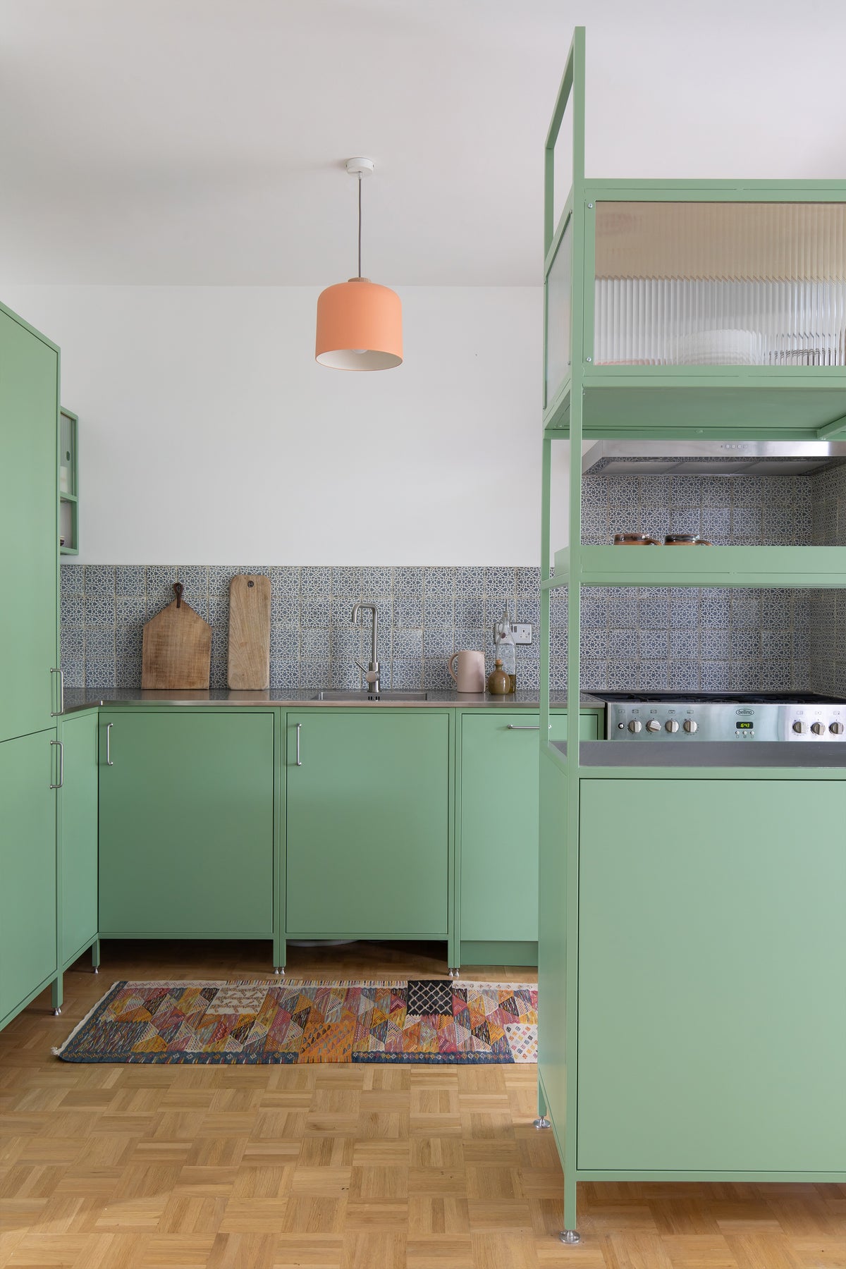 A modern kitchen setup with sage green cabinets and a matching metal shelving unit, featuring a gray patterned tile backsplash, a peach-colored pendant light, and a colorful geometric rug on a parquet wood floor.