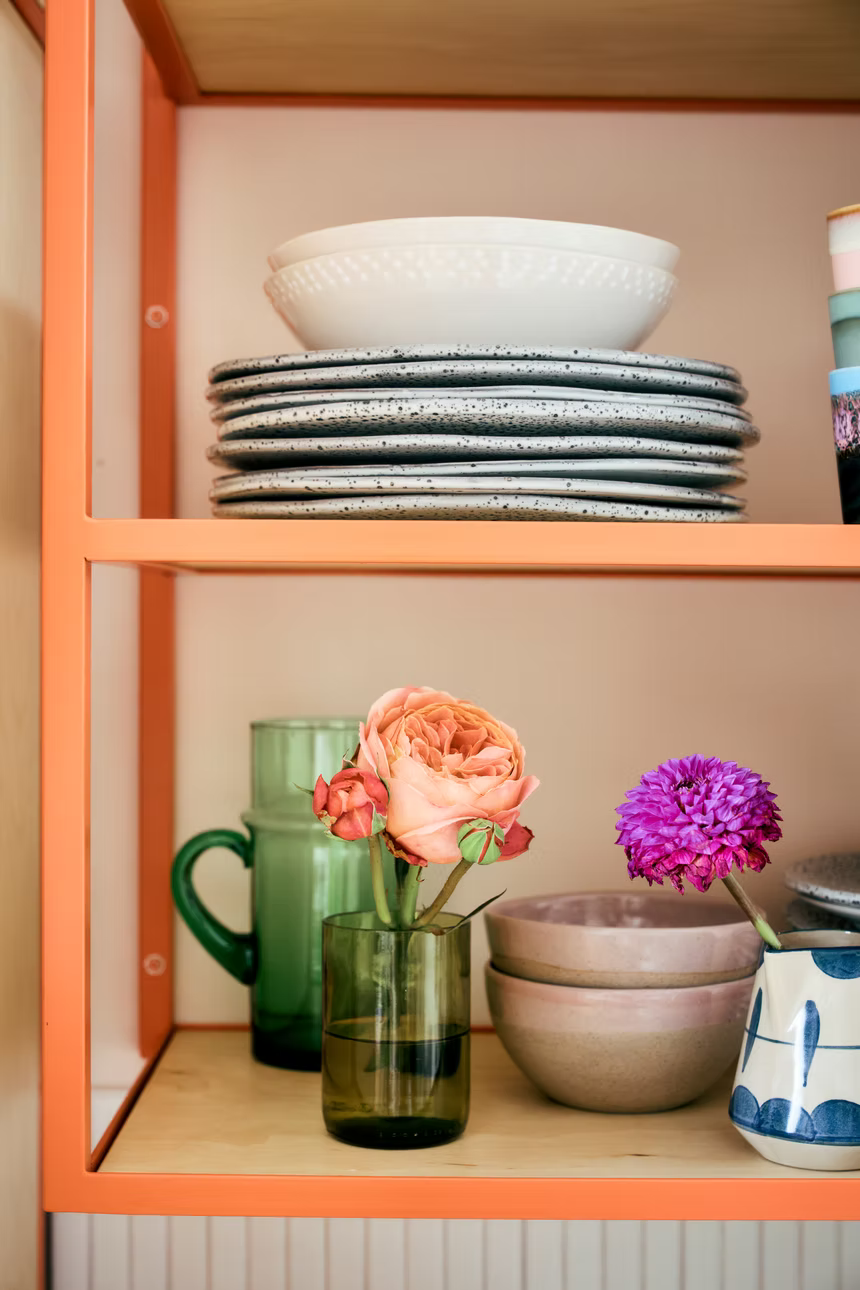 A styled shelf display featuring a stack of speckled gray ceramic plates on the top shelf and a stack of pinkish-beige bowls on the bottom shelf, decorated with a peach rose in a green glass vase and a purple flower in a white and blue patterned pot.