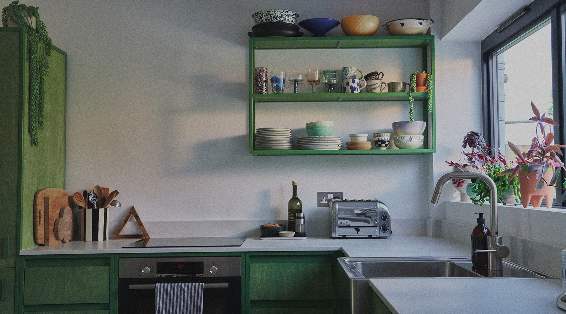 A modern kitchen interior with green cabinets and open shelving holding ceramic dishes, mugs, and bowls, set against white walls and a window with potted plants.