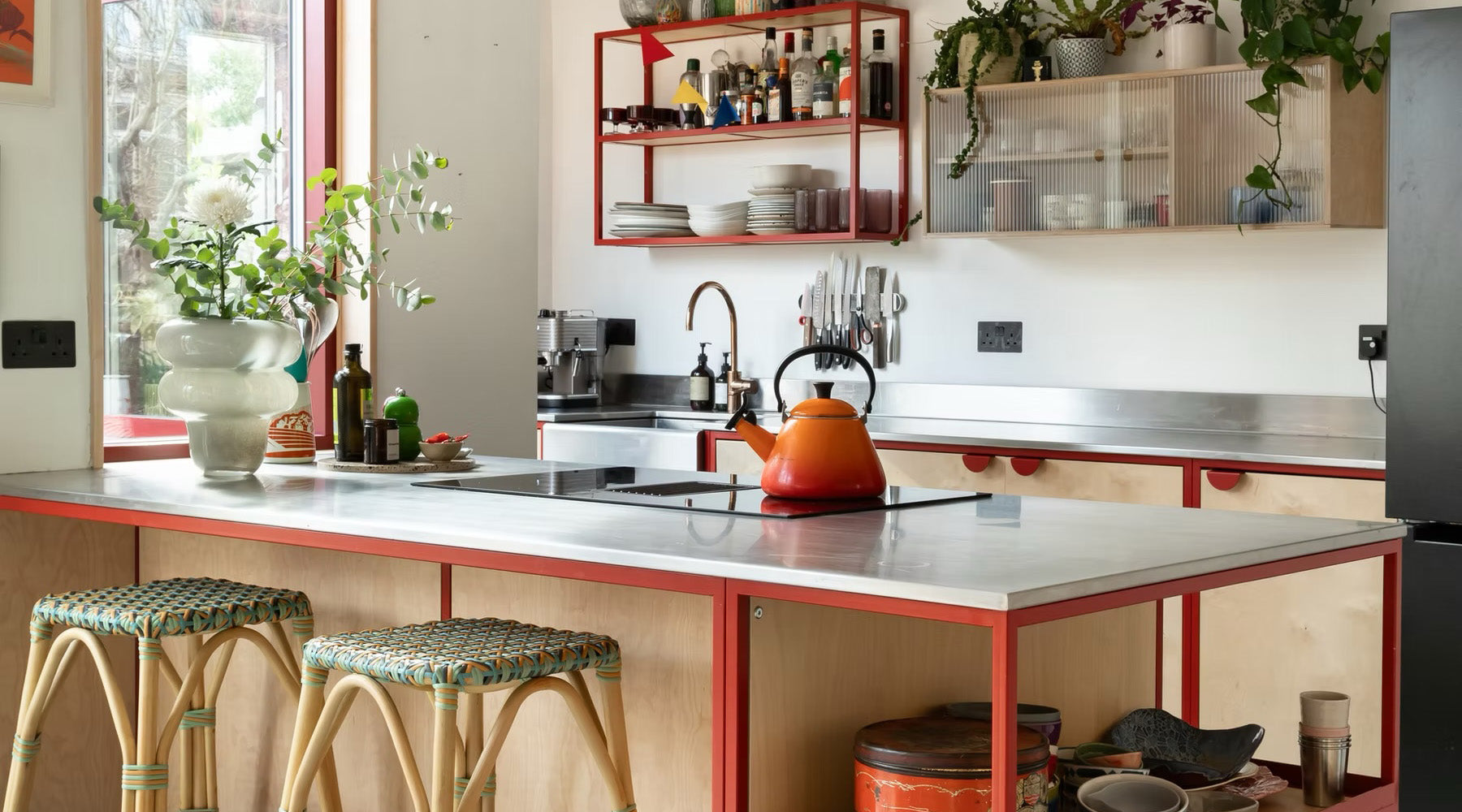 A modern kitchen island with a red metal frame and a light gray countertop, featuring woven bar stools, an orange kettle, and a built-in cooktop. The background shows a white wall with red-framed shelves and cabinets.