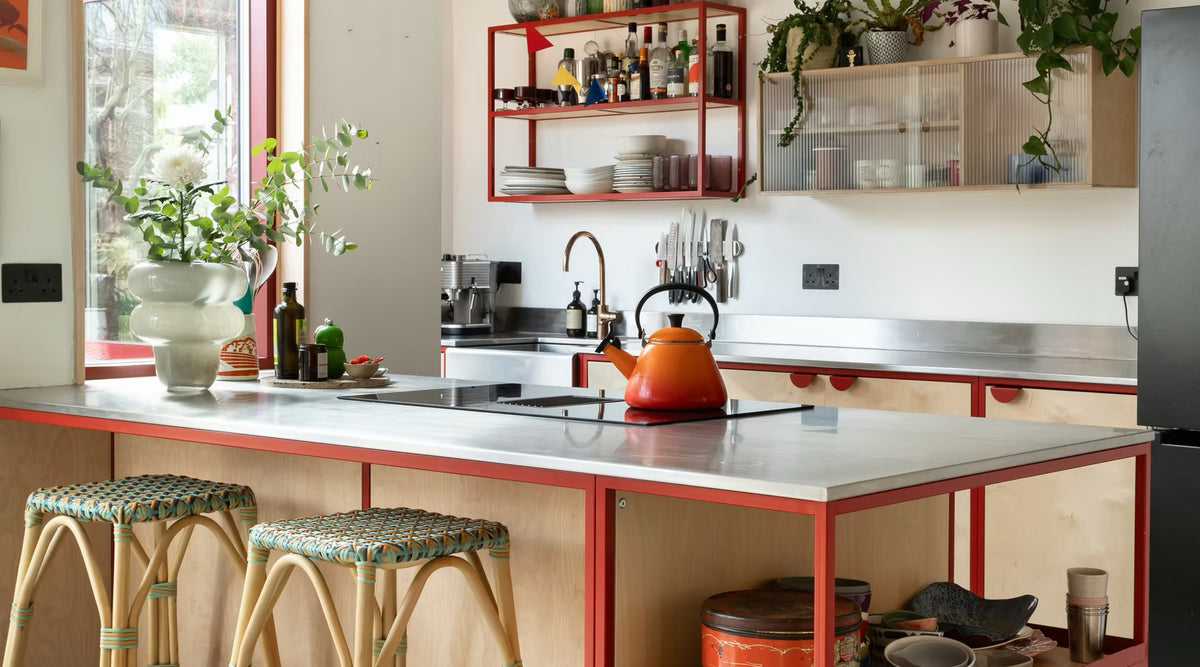 A modern kitchen island with a red metal frame and a light gray countertop, featuring woven bar stools, an orange kettle, and a built-in cooktop. The background shows a white wall with red-framed shelves and cabinets.