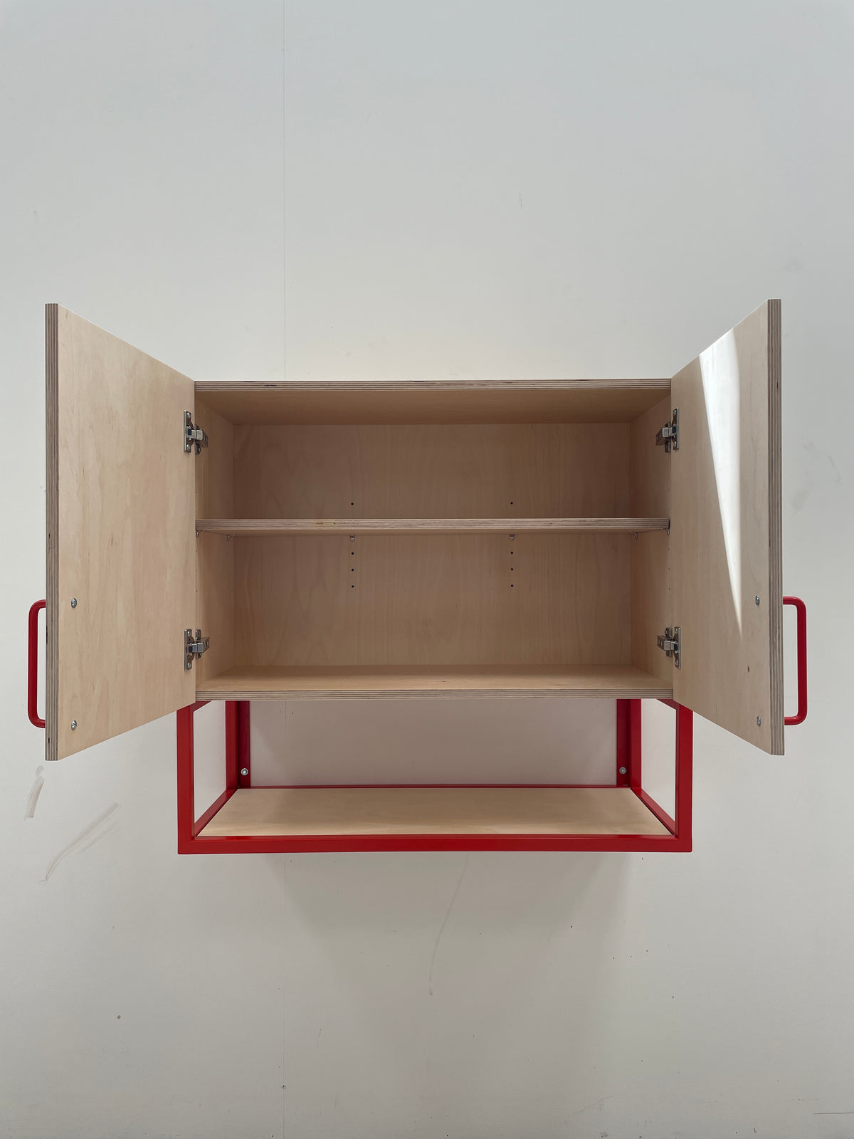 A wall-mounted wooden cabinet with two doors open, showing two interior shelves. The cabinet has a light natural birch plywood finish and a red metal frame with red side handles, mounted against a plain white wall.