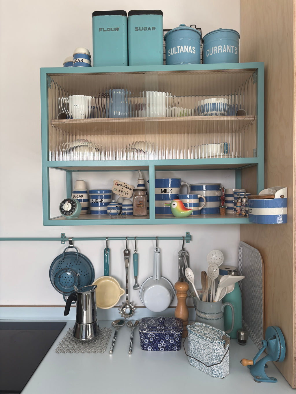 A styled kitchen scene featuring a teal blue open shelving unit mounted on a wall, filled with blue and white canisters, mugs, and glassware. Below the shelves, kitchen utensils hang from a matching teal rack on a white countertop.