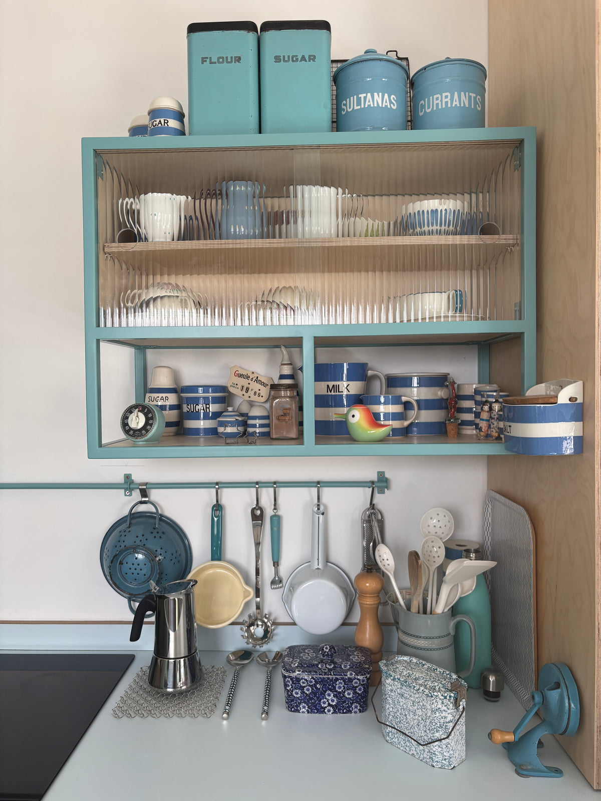 A styled kitchen scene featuring a teal blue open shelving unit mounted on a wall, filled with blue and white canisters, mugs, and glassware. Below the shelves, kitchen utensils hang from a matching teal rack on a white countertop.