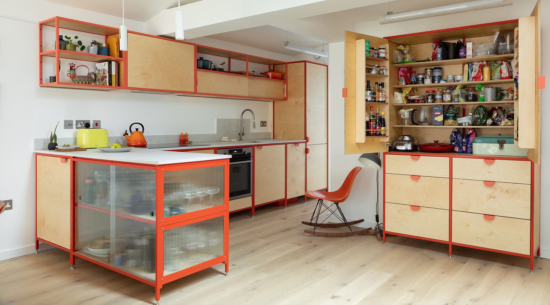 A compact kitchenette with light wood cabinets and bright orange-red metal frames, including a central island with glass doors, wall-mounted cabinets, open shelving, and a small orange rocking chair on a light wood floor.