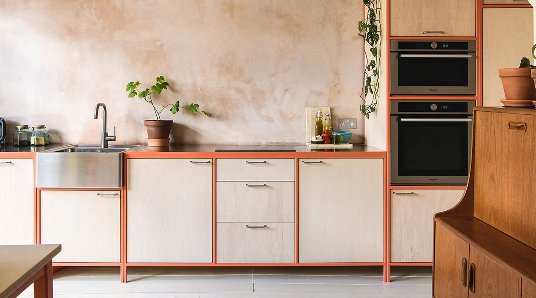 A modern kitchen cabinet setup with light beige wood-grain doors, orange-brown trim, stainless steel sink and appliances, and a textured warm beige wall.