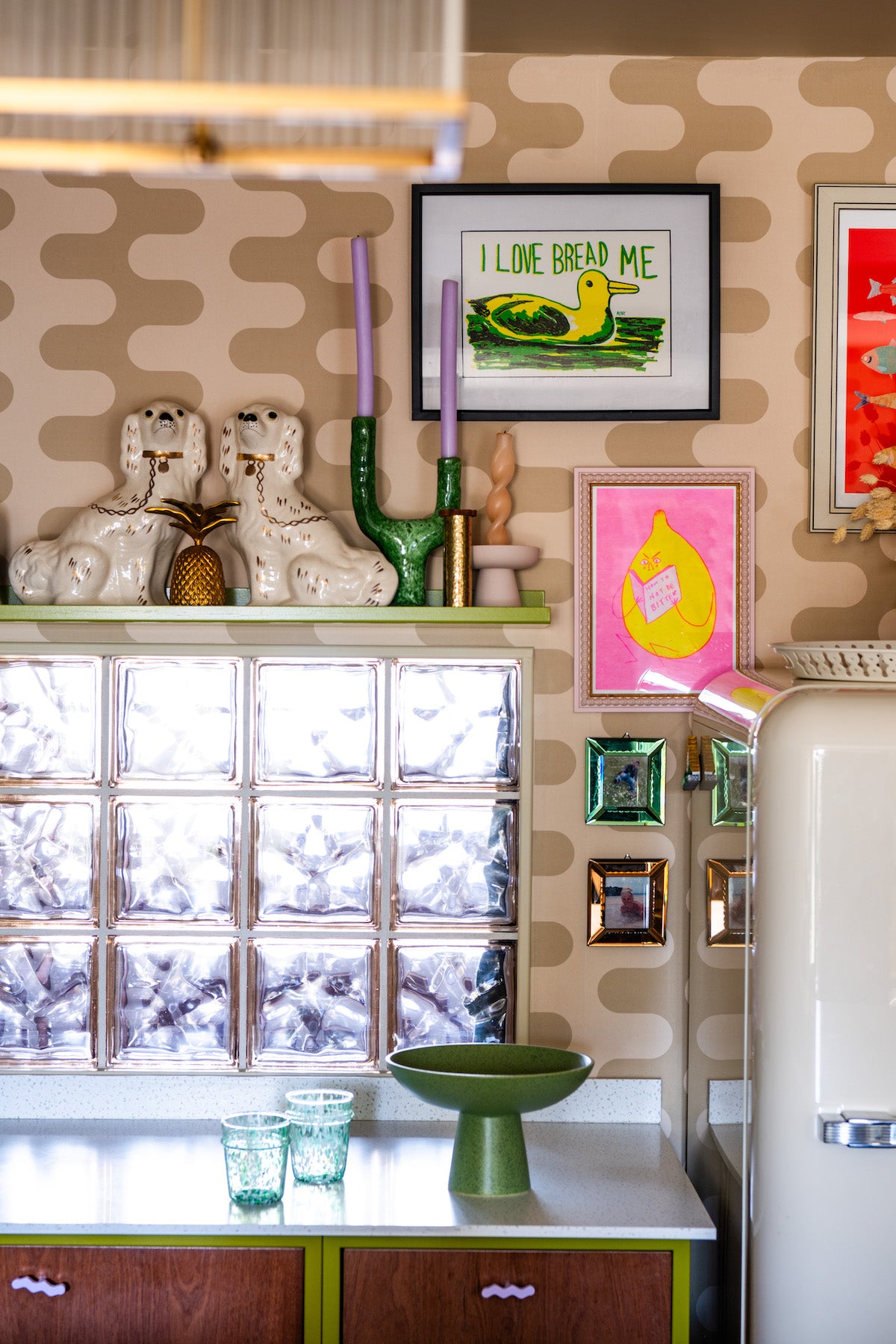 Olive green metal frame kitchen with Sapele fronts, a Smeg fridge, and a solid surface white worktop, set against poodle-print wallpaper in a blonde colourway, with a floating shelf of vintage antiques and knickknacks.
